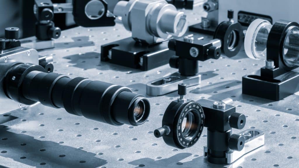 Optical equipment including telescope and lenses are displayed on a table in a laboratory. Optics and photonics concept science experiment with laser background