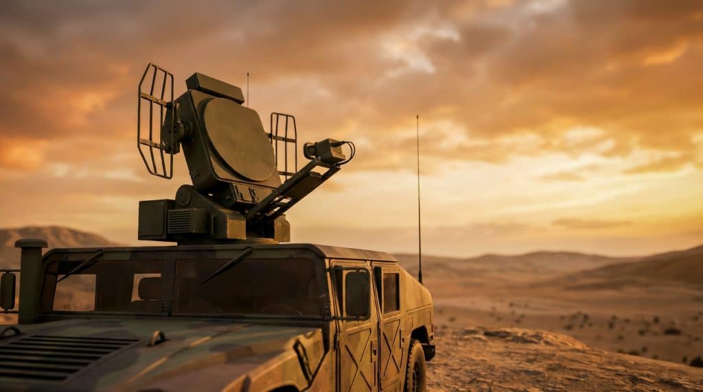 A military radar system is mounted on an armored vehicle in a desert setting. The sky is orange as the sun sets. The focus is on the radar's features and the vehicle's design